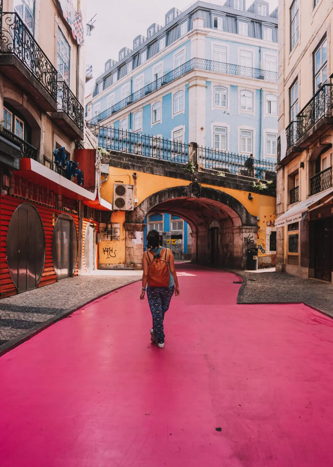 tra i quartieri di lisbona, ecco la pink street: Io nel mezzo di una strada rosa, sormontata da un ponticello pedonale giallo, e sullo sfondo, un palazzo azzurro